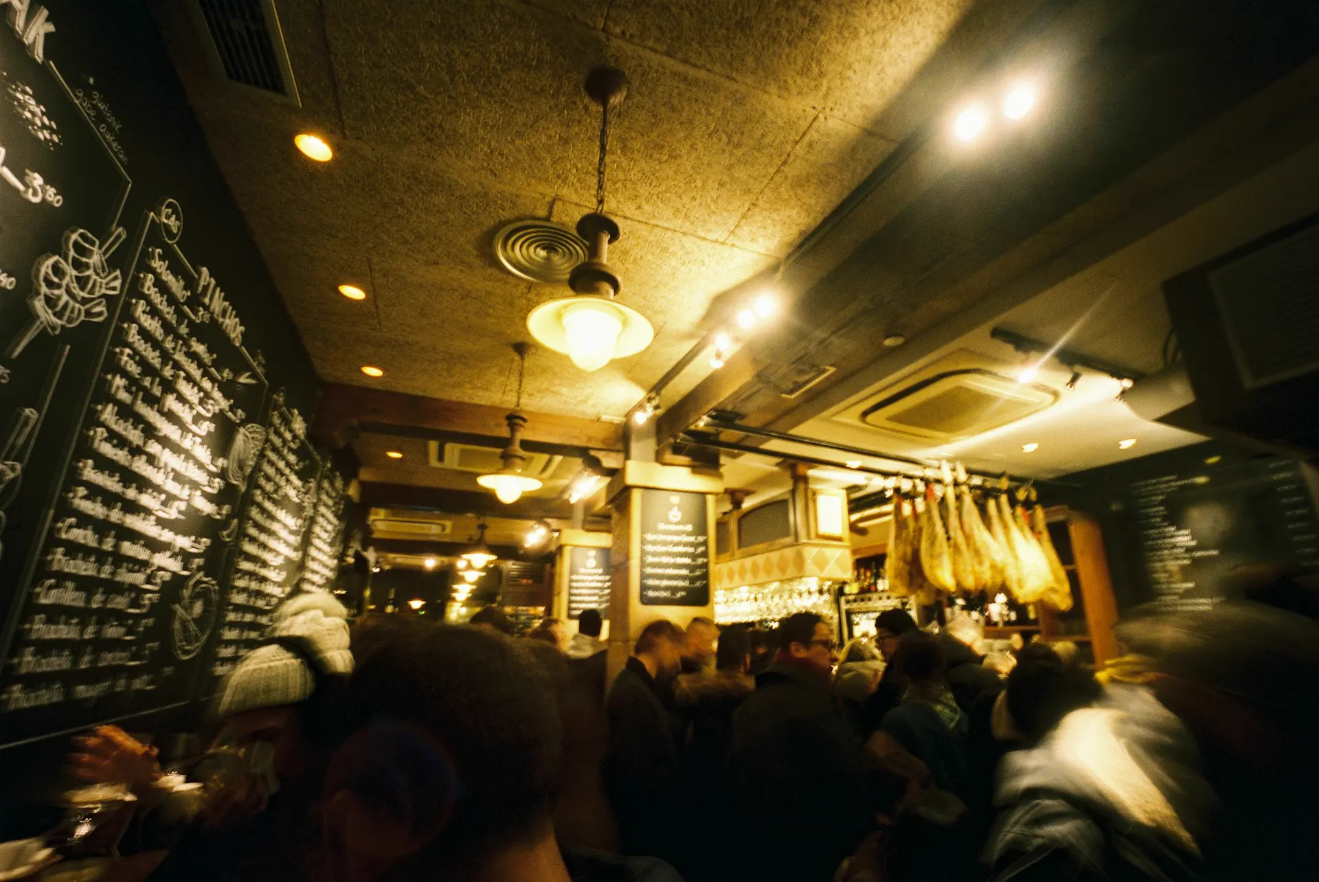Una barra de pintxos llena de gente, con jamones colgados y luz cálida